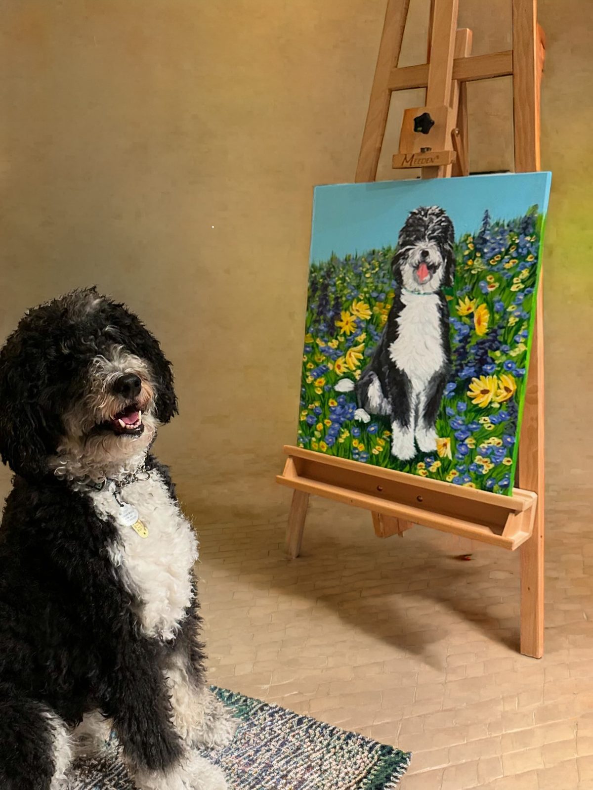 Zoya, a black-and-white Bernedoodle, sits beside an easel holding her painted portrait in a field of blue and yellow wildflowers
